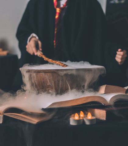 Osoba w magicznej szacie przeprowadzająca rytuał nad dymiącym naczyniem, otoczona przez otwarte księgi i zapalone świeczki, z tablicą zaklęć w tle, co tworzy atmosferę tajemniczości, magii i kreatywności.