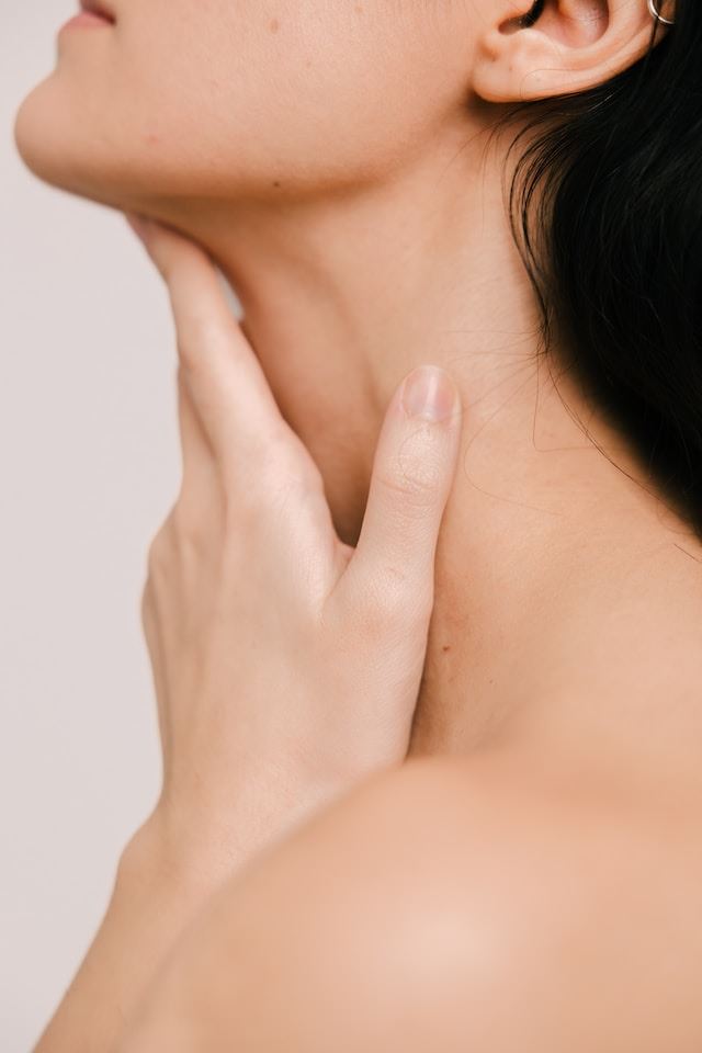 Kobieta dotyka swojej szyi z troską, wykonuje samobadanie tarczycy.