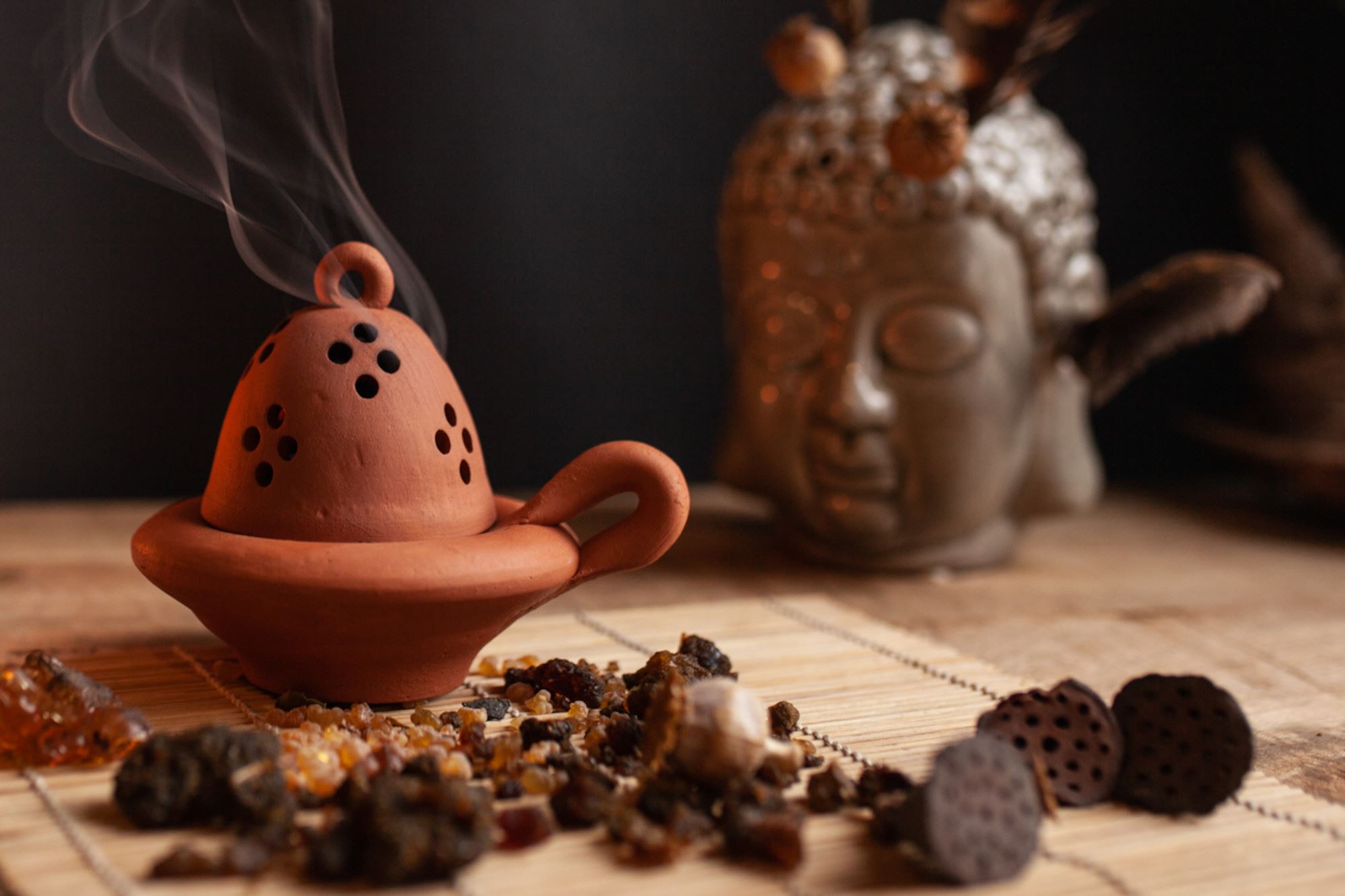 Gliniana kadzielniczka z której unosi się dym, obok kadzidła, w tle figurka głowy buddy.
